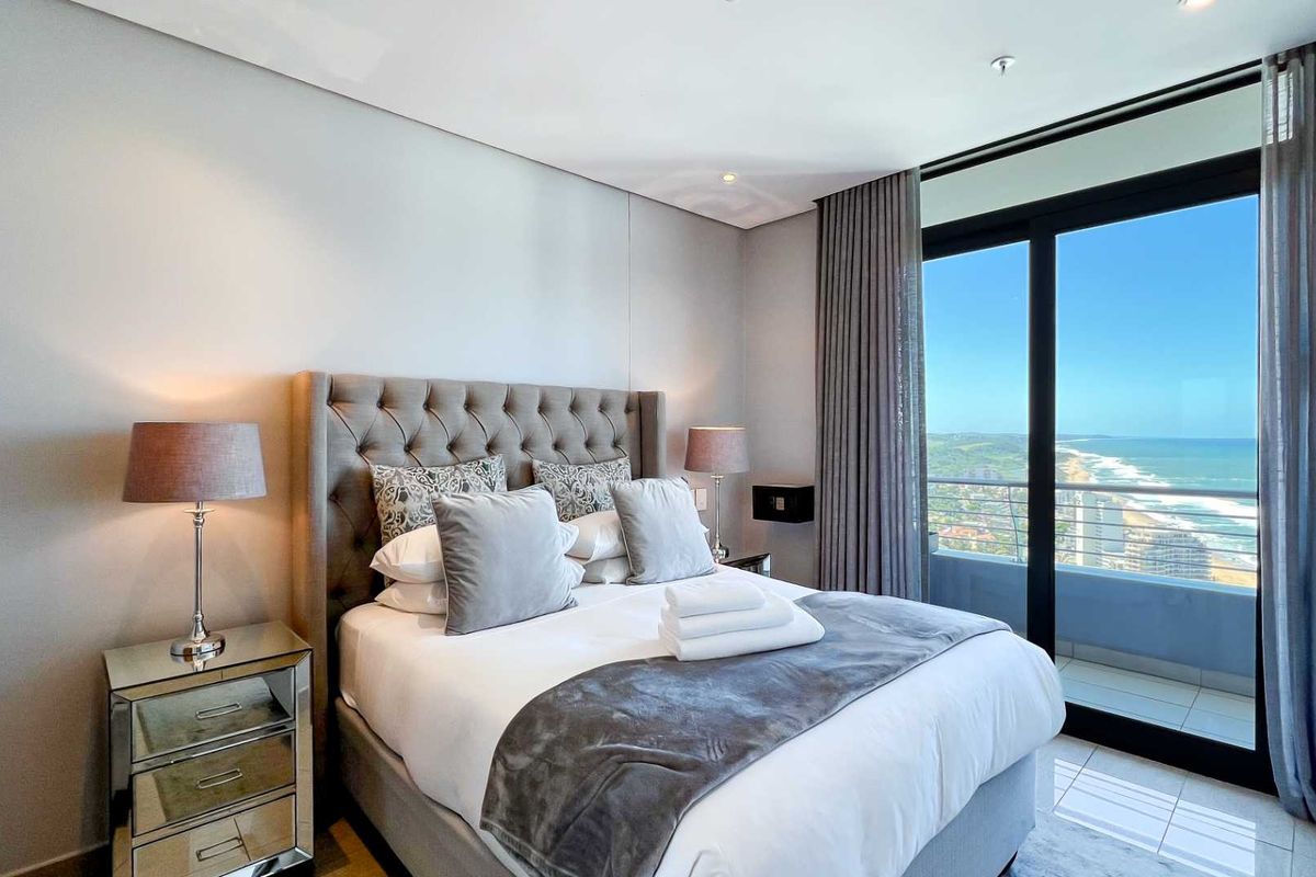 Modern bedroom featuring neutral finishes, recessed lighting, and large sliding glass doors opening to a balcony with expansive coastal views.