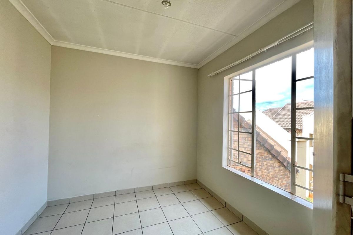 A bright, empty bedroom featuring neutral tiled flooring, a large window with metal burglar bars, and a simple ceiling light fixture.