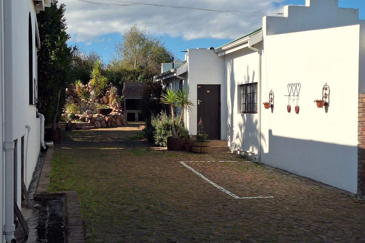 A rear exterior view featuring a brick-paved courtyard, a built-in braai area, and a landscaped rockery garden.
