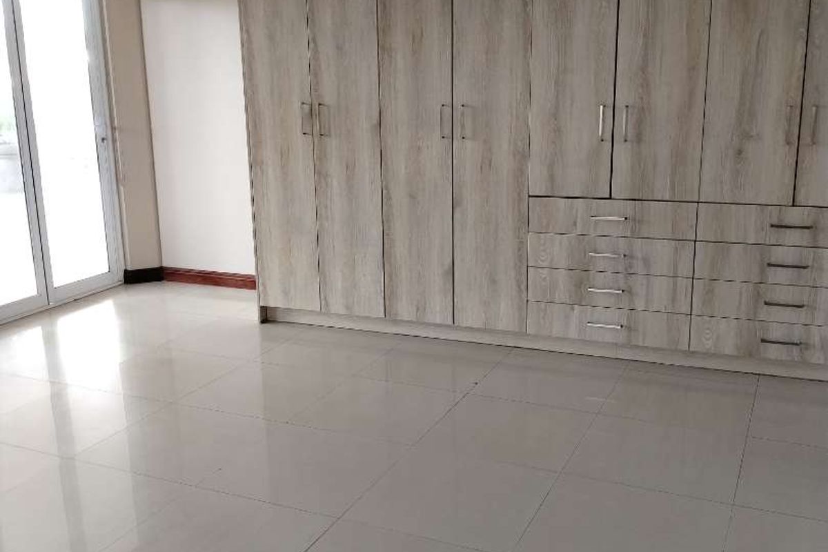 A bright, empty bedroom featuring extensive built-in wooden cupboards, polished tiled flooring, and a sliding glass door providing natural light.