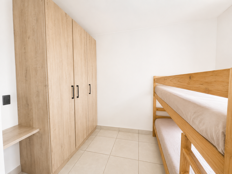 A clean bedroom featuring light wood-toned built-in wardrobes, a matching built-in desk, and neutral tiled flooring.