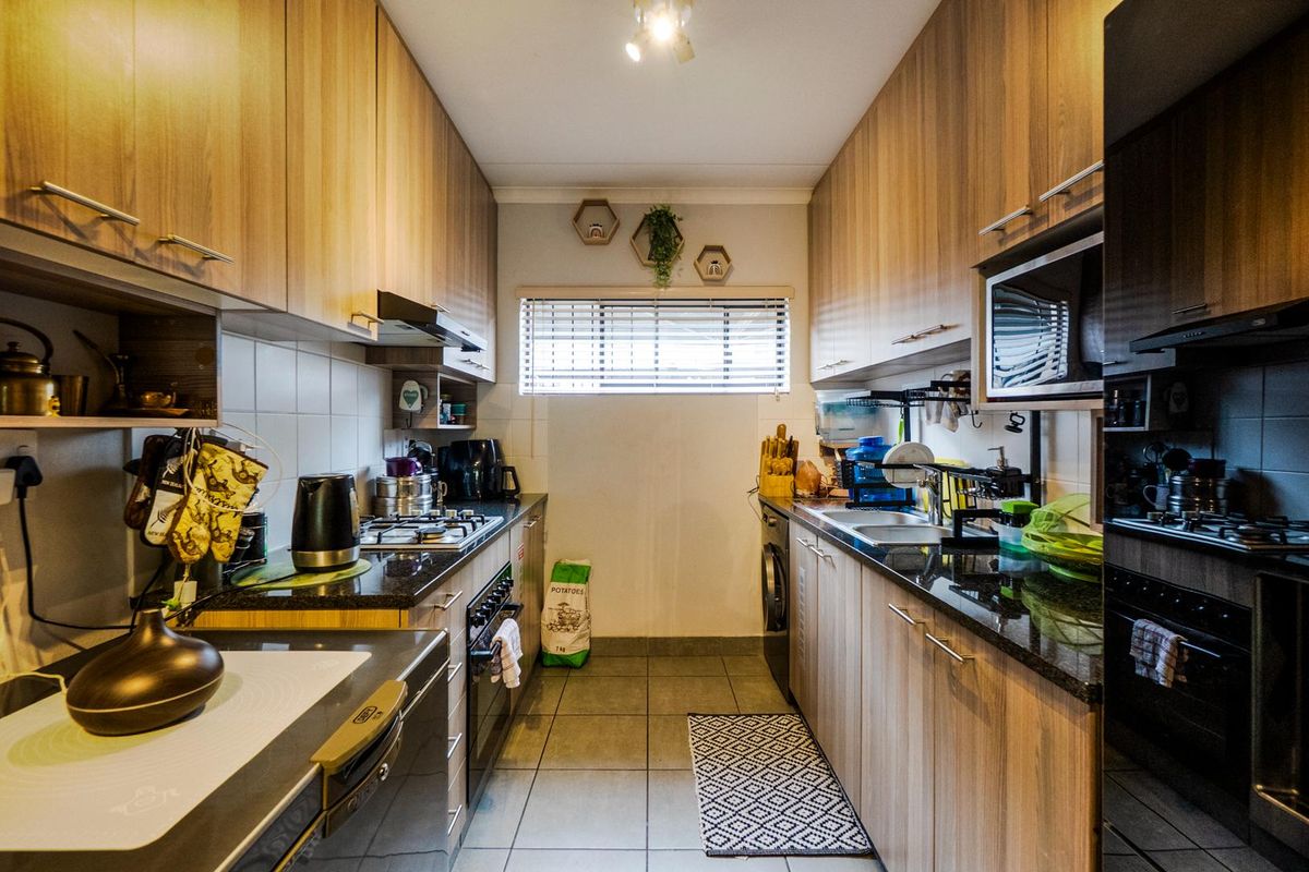 A galley-style kitchen featuring wood-look cabinetry, granite countertops, a built-in oven, gas hob, extractor fan, and integrated laundry plumbing.