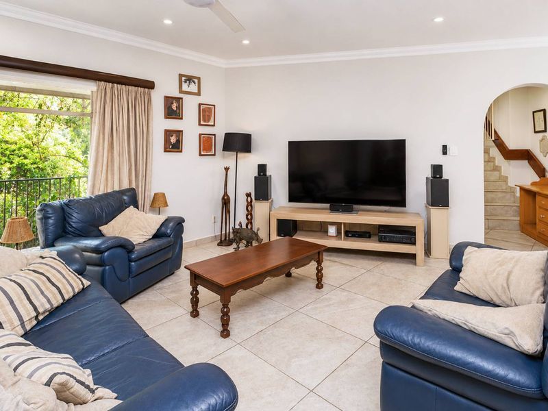 Spacious living room featuring neutral tiled flooring, recessed lighting, and an arched entryway leading to a staircase, with access to a balcony.
