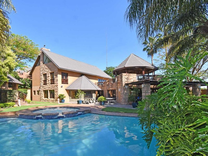 A well-maintained outdoor pool area featuring a swimming pool, stone-clad building exterior, covered patio, and a gazebo set within a landscaped garden.