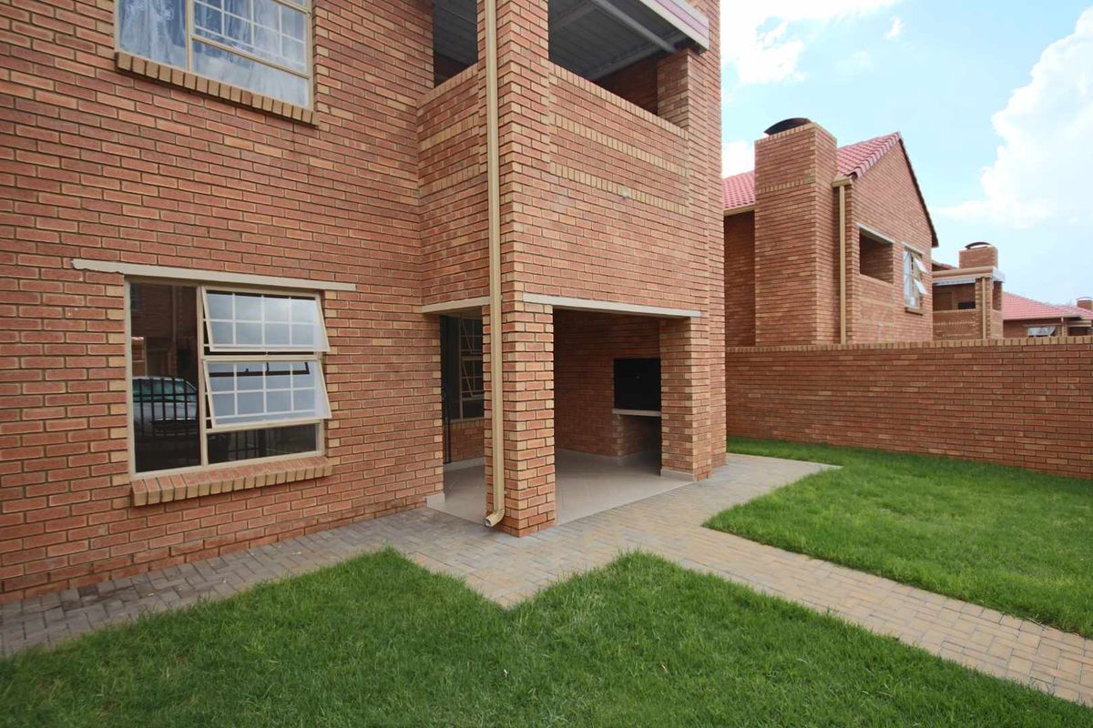 Rear view of the apartment featuring a brick facade, a covered patio with a built-in braai, a paved walkway, and a private lawn area.