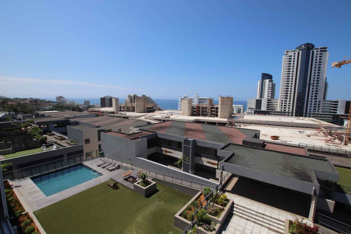 Elevated aerial view of the apartment complex featuring a swimming pool, landscaped garden areas, and surrounding urban development with distant ocean views.