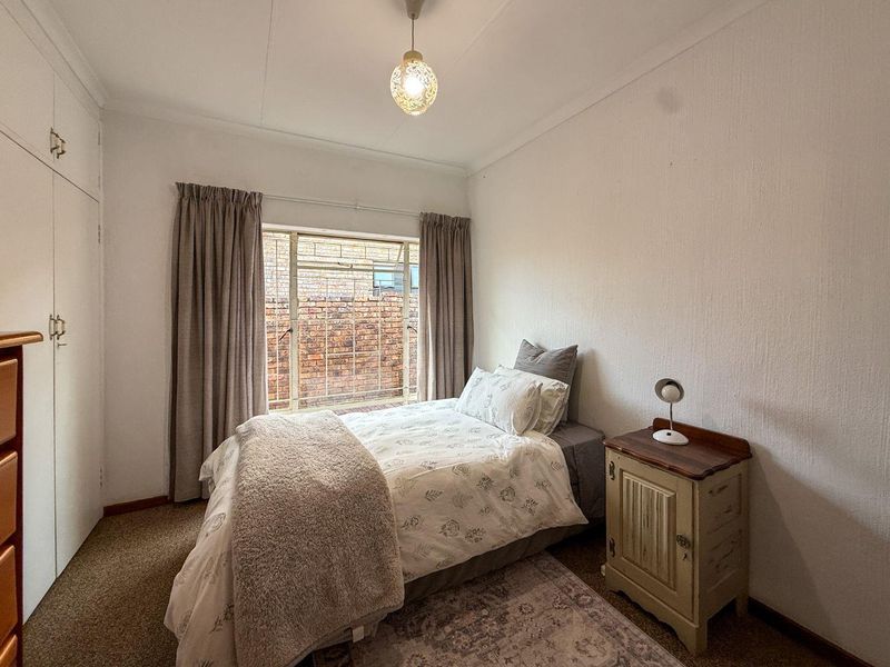 A functional bedroom featuring built-in cupboards, carpeted flooring, and a steel-framed window overlooking a brick wall.