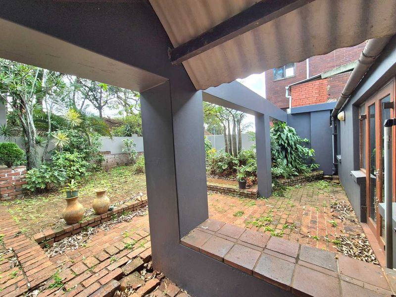 A covered outdoor patio area featuring brick paving and direct access to the home through timber-framed glass doors, overlooking a garden space.