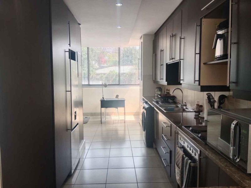 A galley-style kitchen featuring dark cabinetry, integrated appliances, tiled flooring, and a bright transition area leading to a windowed balcony space.