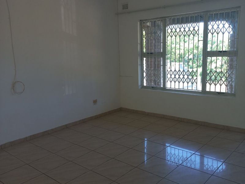 A bright, empty living area featuring neutral tiled flooring and a large window fitted with a security gate.