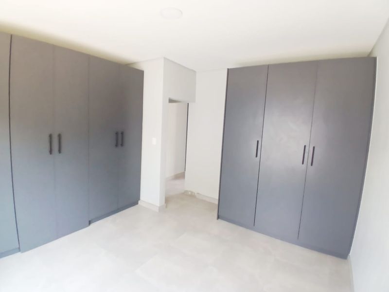 A bright, modern bedroom featuring extensive built-in grey cabinetry and neutral tiled flooring, offering a clean and spacious layout.