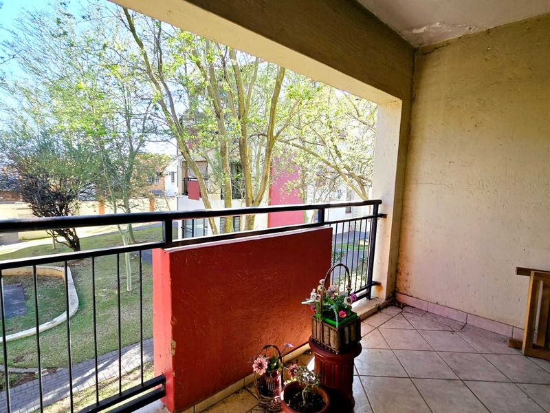 A covered balcony featuring tiled flooring and a metal balustrade, overlooking a communal garden area. The space requires maintenance to the walls and ceiling.