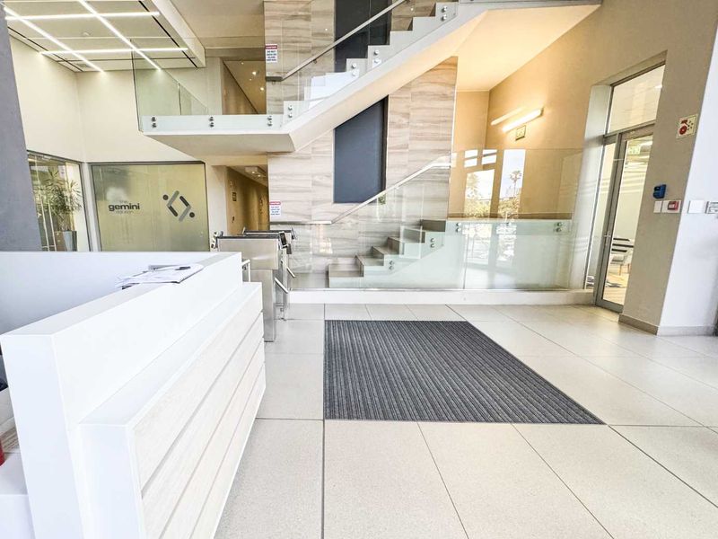 Modern commercial building entrance featuring a reception desk, glass staircase with balustrades, security turnstiles, and polished tiled flooring.