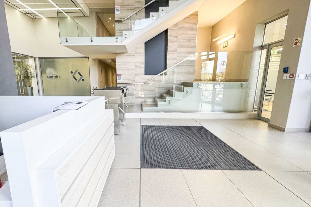 Modern commercial building entrance featuring a reception desk, glass staircase with balustrades, security turnstiles, and polished tiled flooring.