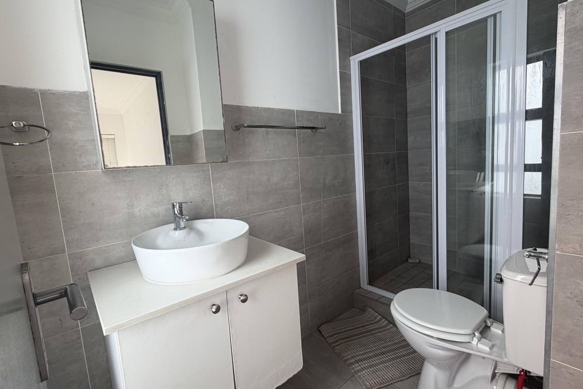 A functional bathroom featuring grey wall tiles, a modern vessel sink on a vanity cabinet, a toilet, and a glass-enclosed shower stall.