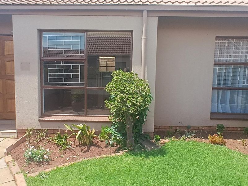 Front facade of the townhouse featuring a paved entrance, timber door, aluminium windows with security bars, and a well-maintained garden area.