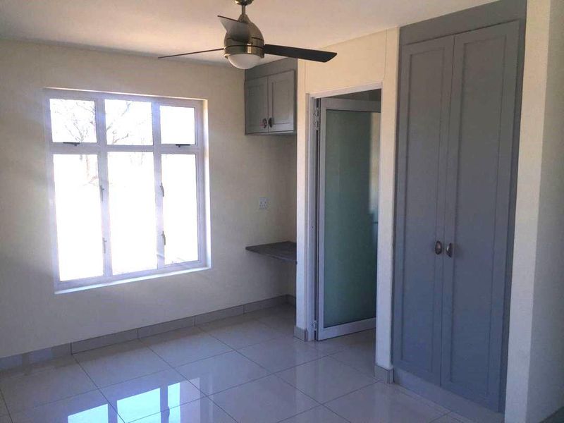 A bright, empty bedroom featuring tiled flooring, a ceiling fan, built-in cupboards, and a convenient built-in desk area.
