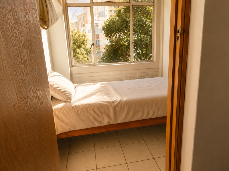 A compact bedroom featuring tiled flooring, a large window providing natural light, and painted brick wall detailing.