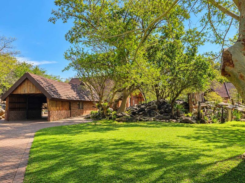 A well-maintained exterior view of a property featuring a brick driveway, landscaped garden with mature trees, and a building with a thatched roof.