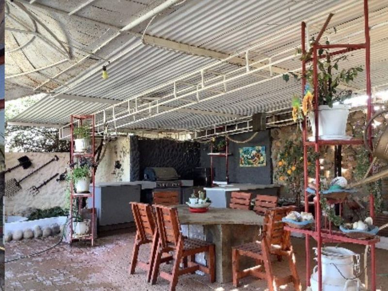 A covered outdoor patio area featuring a built-in braai, stone wall accents, and a corrugated metal roof, suitable for farm-style outdoor entertaining.