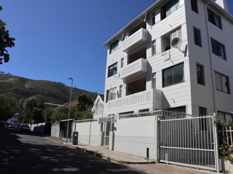 A multi-story apartment building featuring a secure perimeter wall, gated entrance, and private balconies, situated on a paved street.
