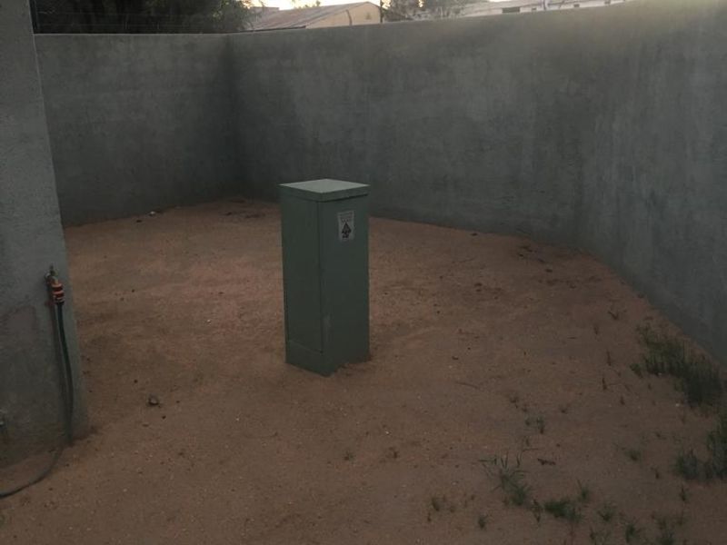 A small, enclosed rear outdoor area featuring a perimeter wall, an outdoor tap, and a central electrical utility box on an unpaved surface.
