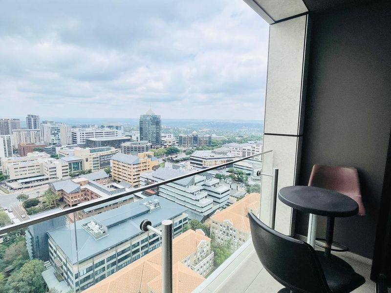 A modern apartment balcony featuring a glass balustrade and stainless steel handrail, offering expansive elevated views of the surrounding city skyline.
