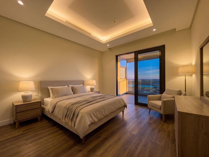 A modern, well-lit bedroom featuring a recessed tray ceiling with ambient cove lighting, wood-look flooring, and direct access to a private balcony.