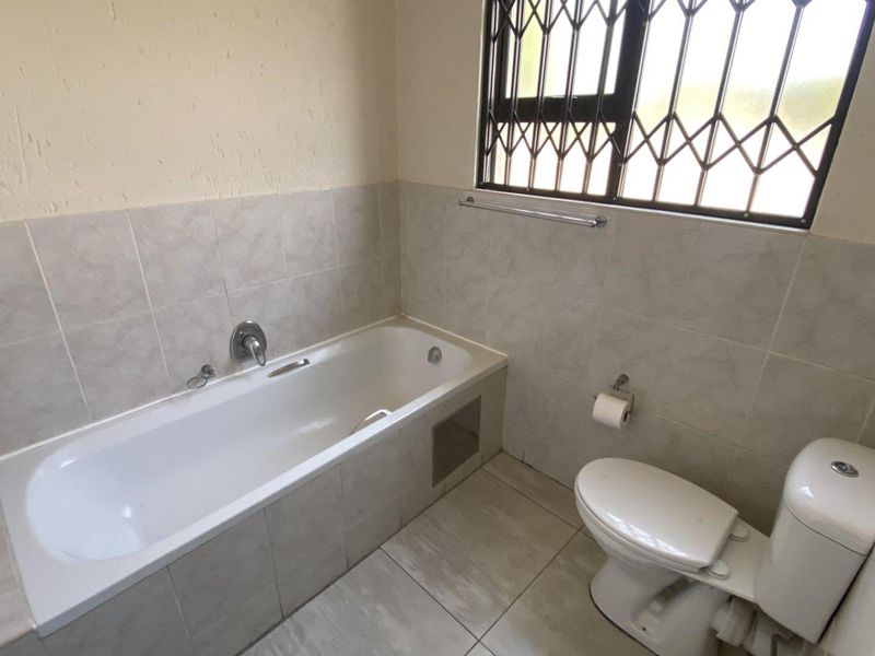 A functional bathroom featuring a built-in bathtub, toilet, and neutral wall and floor tiling, complete with a security gate on the window.