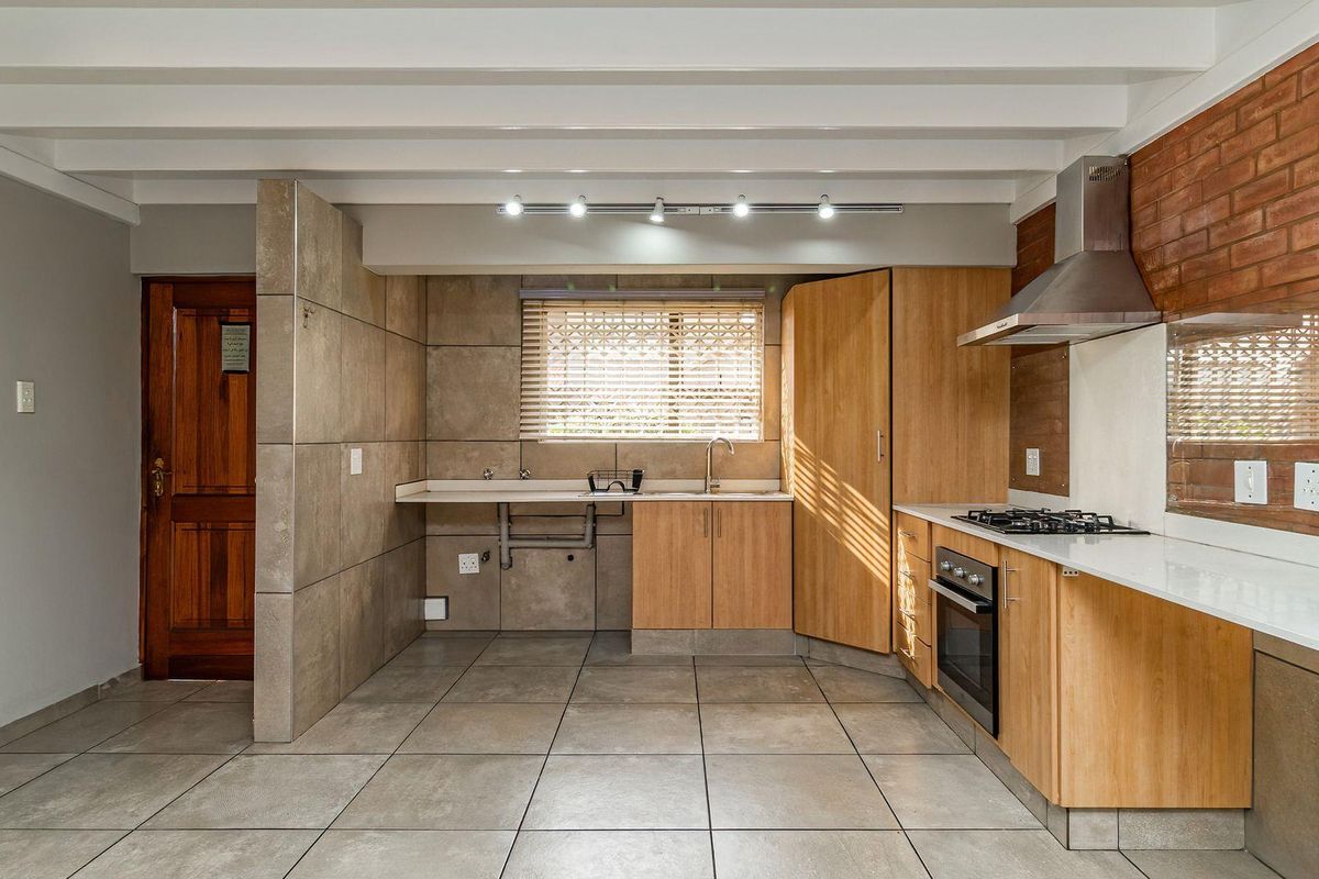 A modern kitchen featuring light wood cabinetry, stone countertops, a gas hob, built-in oven, and a distinctive exposed brick feature wall.
