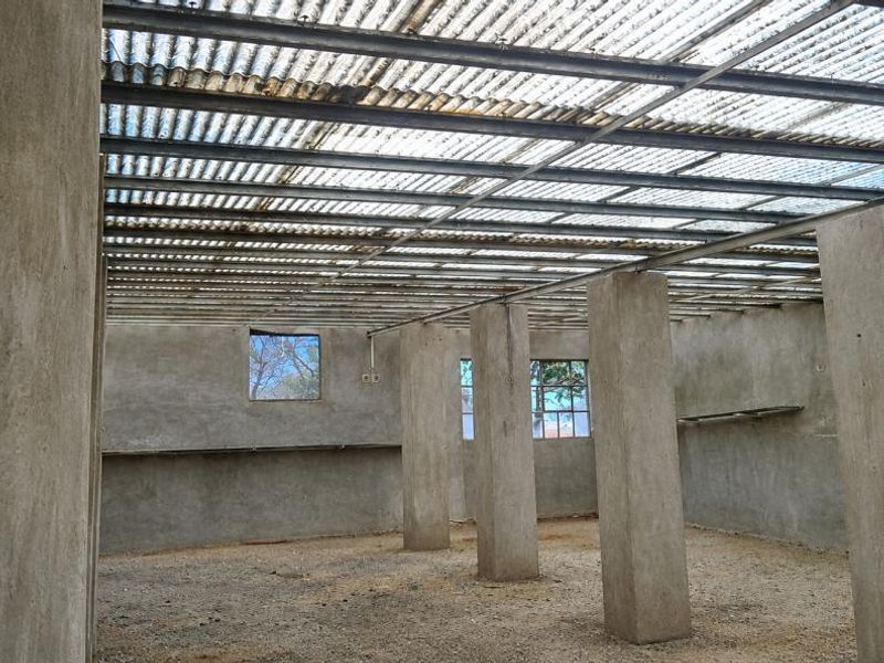 An unfinished utility space featuring concrete columns, exposed steel roof structure with translucent sheeting, and raw cement plastered walls.