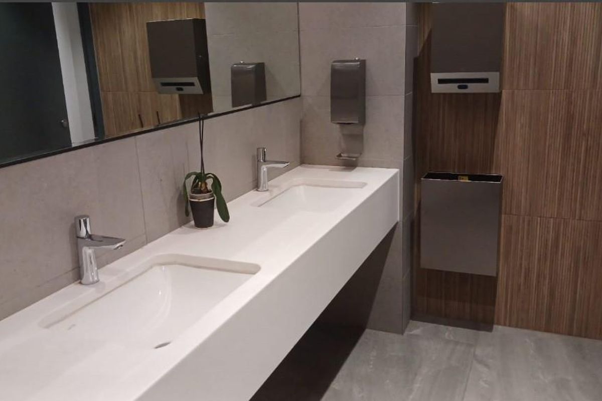 Modern office bathroom featuring a sleek double vanity with integrated sinks, contemporary wall-mounted faucets, and a combination of grey tile and wood-look wall finishes.