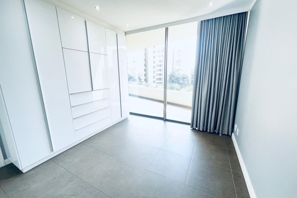 A bright, modern bedroom featuring sleek white built-in cupboards, large format floor tiles, and a sliding glass door leading to a balcony.