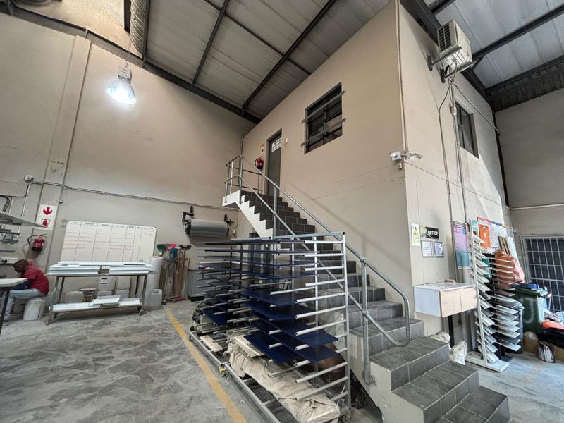 Industrial warehouse space featuring high ceilings with exposed steel trusses, concrete flooring, and a tiled staircase leading to an elevated office area.