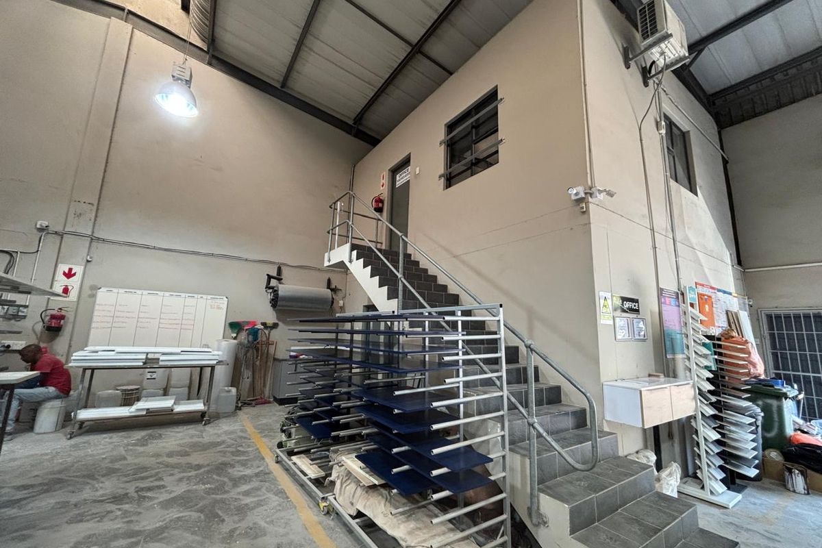 Industrial warehouse space featuring high ceilings with exposed steel trusses, concrete flooring, and a tiled staircase leading to an elevated office area.