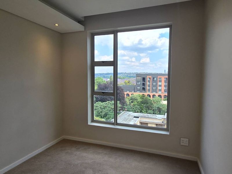 A bright, empty bedroom featuring neutral walls, carpeted flooring, recessed ceiling lighting, and a large window offering an urban view.