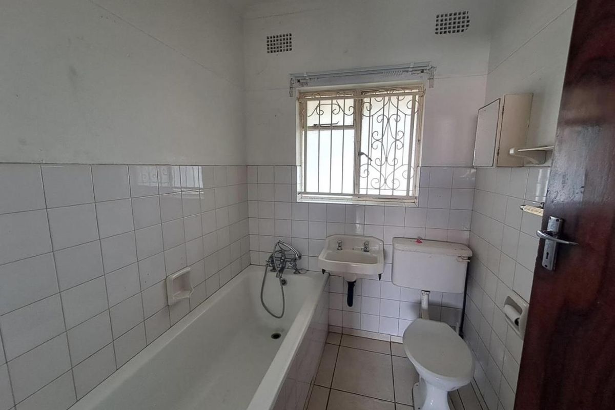 A dated bathroom featuring a standard bathtub, wall-mounted basin, and toilet, with white wall tiling and a security-grilled window.