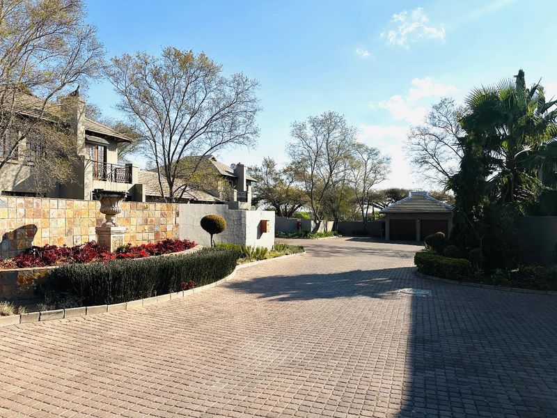 A well-maintained townhouse complex entrance featuring a paved driveway, stone-clad retaining walls, landscaped garden beds, and a double garage.