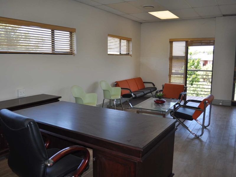 A spacious office area featuring tiled flooring, recessed ceiling lighting, and large windows with venetian blinds, providing natural light and access to a balcony.