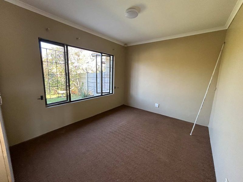 An empty bedroom featuring carpet flooring, neutral wall paint, and a large window with security bars overlooking the garden.