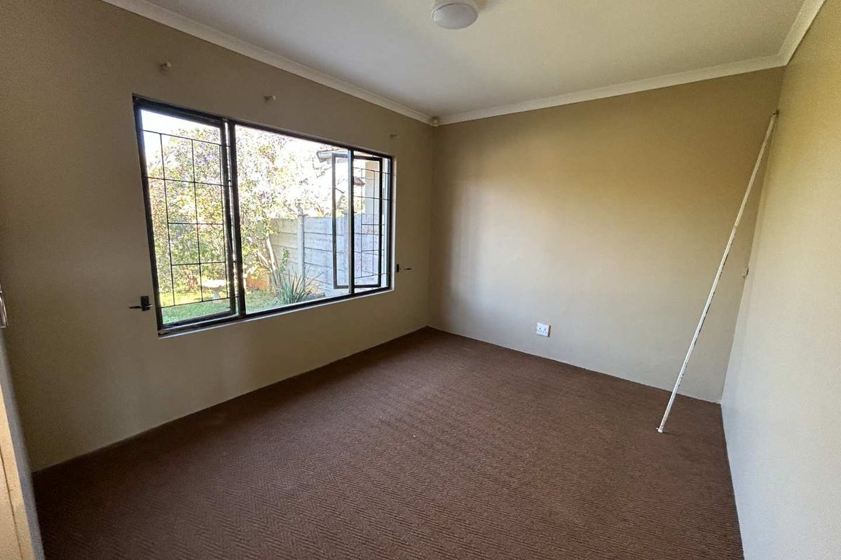 An empty bedroom featuring carpet flooring, neutral wall paint, and a large window with security bars overlooking the garden.