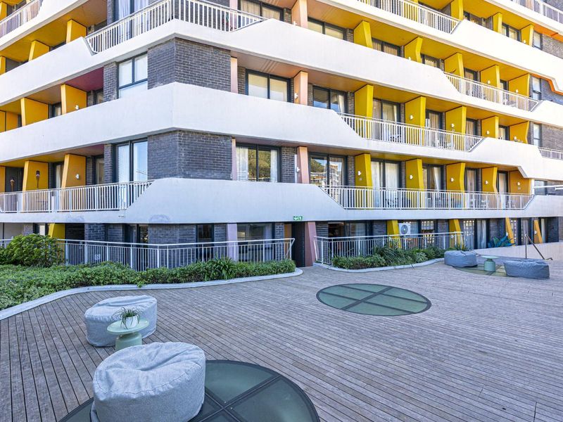 A view of the apartment building exterior featuring a spacious timber deck terrace with integrated glass floor panels and landscaped garden borders.
