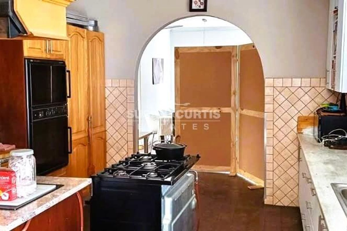 A functional kitchen featuring wooden cabinetry, a built-in oven, a gas hob, and a tiled backsplash, leading through an arched doorway to adjacent living space.