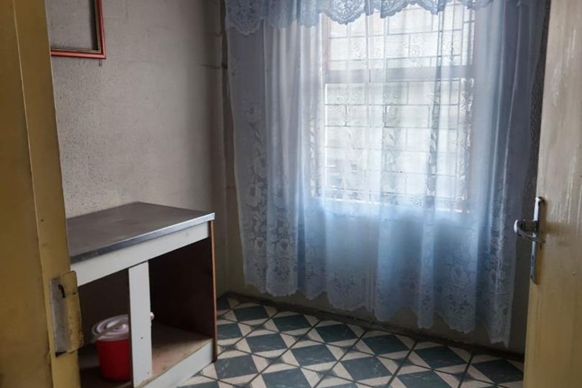 A compact bedroom featuring patterned floor tiles, security bars on the window, and a built-in vanity unit, offering a blank canvas for renovation.