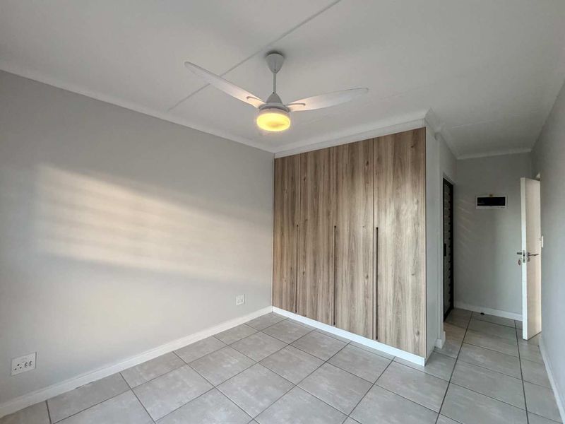 A bright, empty bedroom featuring modern wood-finish built-in cupboards, a ceiling fan, and neutral tiled flooring.