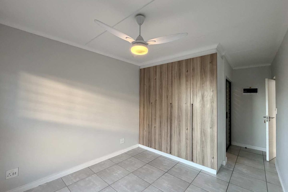 A bright, empty bedroom featuring modern wood-finish built-in cupboards, a ceiling fan, and neutral tiled flooring.