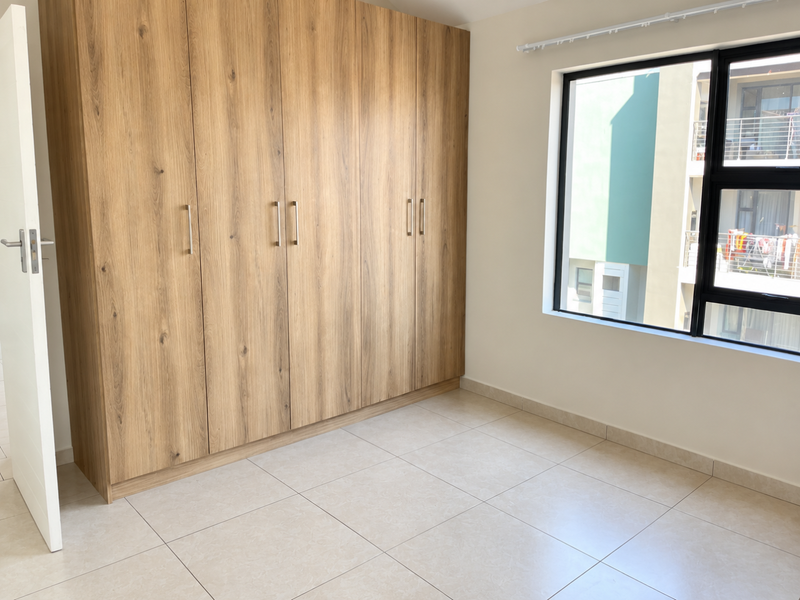 A bright, empty bedroom featuring modern wood-finish built-in cupboards, neutral tiled flooring, and a large window providing natural light.