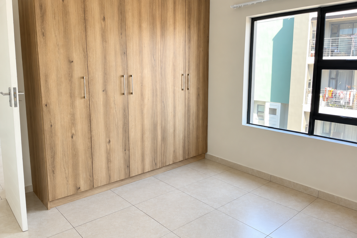 A bright, empty bedroom featuring modern wood-finish built-in cupboards, neutral tiled flooring, and a large window providing natural light.