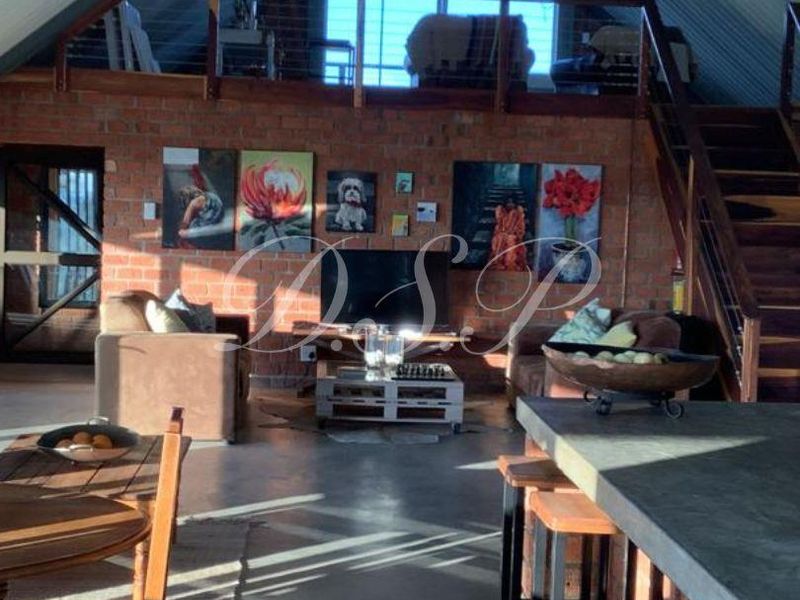 Spacious open-plan living area featuring exposed brick walls, vaulted corrugated metal ceilings, a concrete kitchen island, and a mezzanine level with a timber staircase.