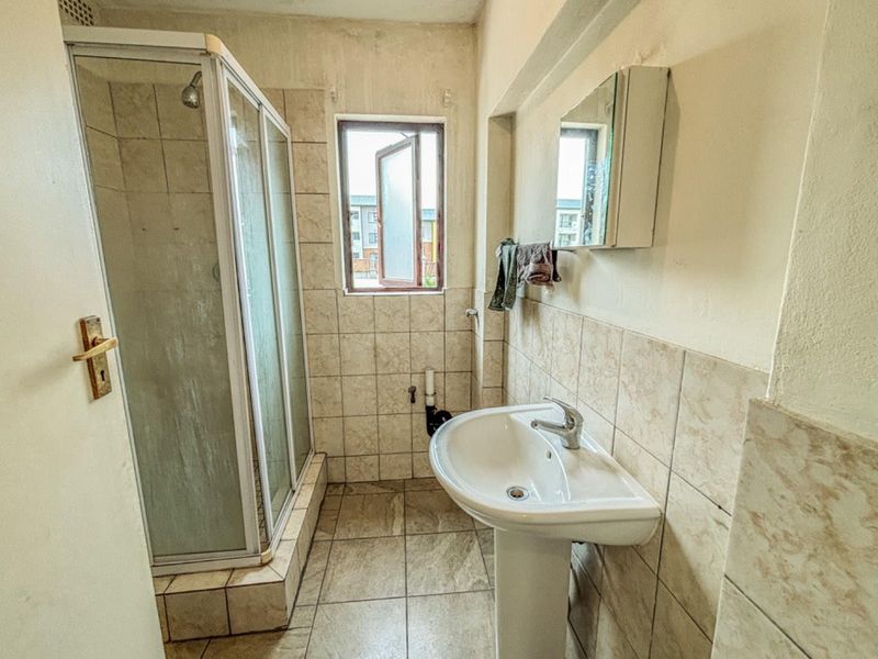 A compact bathroom featuring a pedestal sink, a corner shower enclosure with tiled surround, and a small window for natural ventilation.